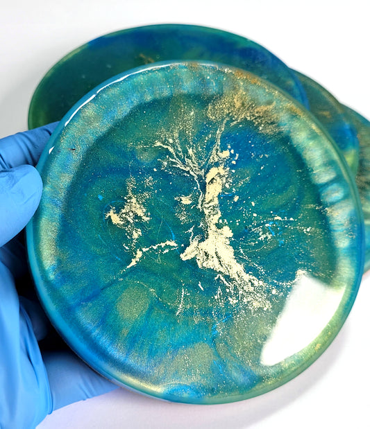 A hand in a blue glove holds a Blue Green and Gold Coaster by The Cerulean Wolf, featuring swirling abstract patterns. Other handmade coasters from the collection are visible underneath.