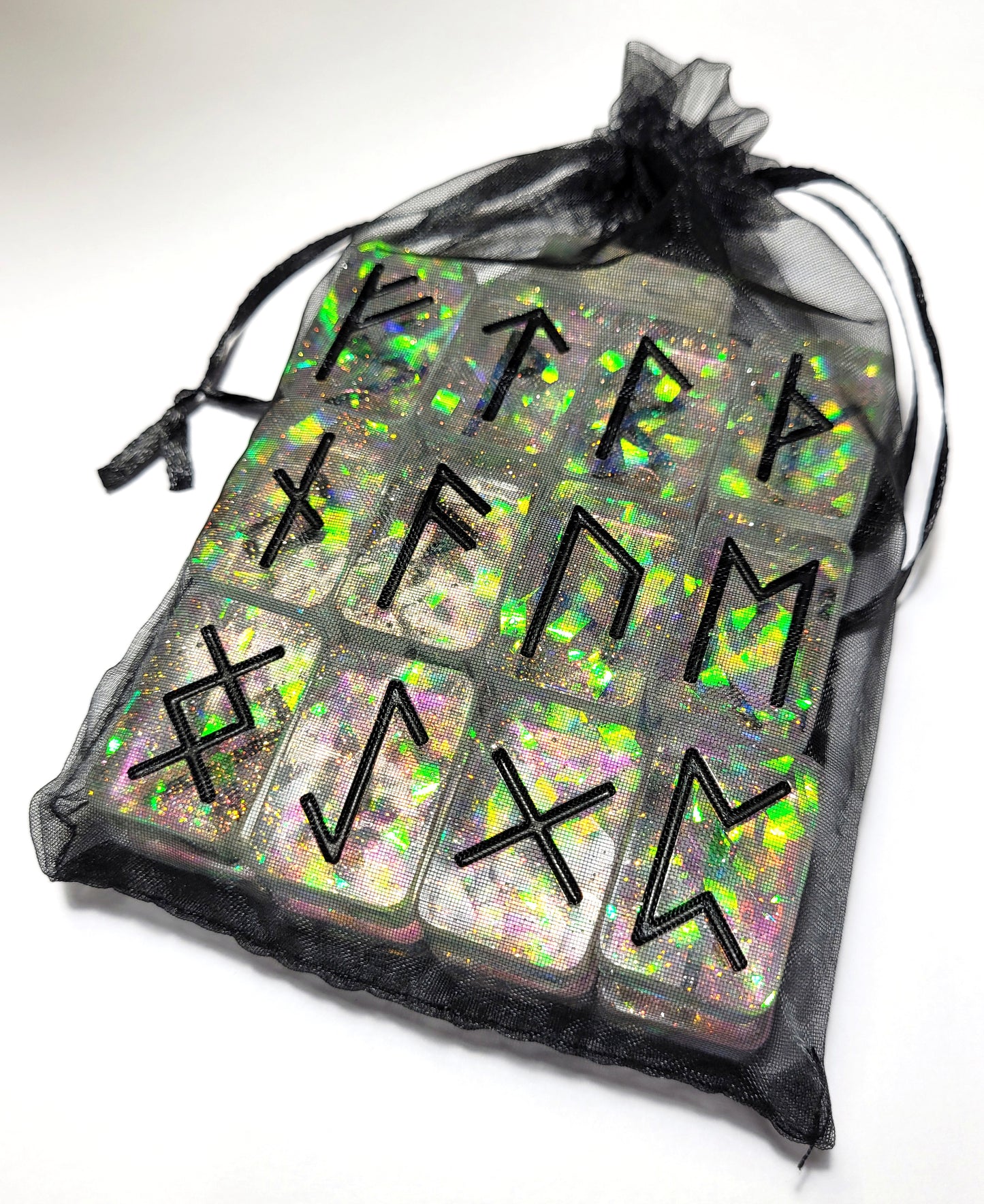 A black mesh drawstring bag holds The Cerulean Wolf’s 25 Holographic and Black Elder Futhark Runes, handmade from epoxy resin with iridescent glitter and black engravings, each arranged in a grid pattern.