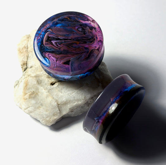 The Cerulean Wolf’s Midnight Mist Double Flared Plugs, featuring swirling purple, blue, and pink epoxy resin, rest on a rough white stone surface under bright lighting.