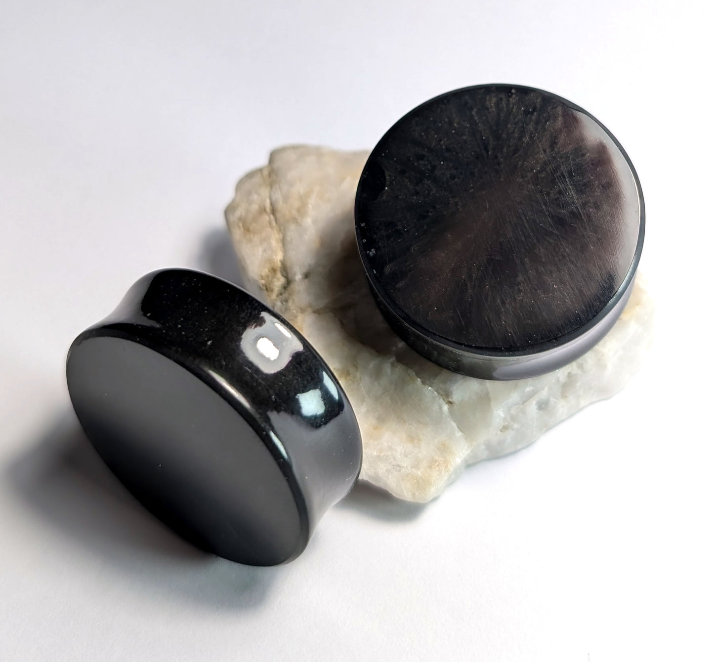 Two round Black Depths Double Flared Plugs by The Cerulean Wolf, with a polished, glossy finish and reflective surfaces, rest on a rough white stone surface against a clean white background.