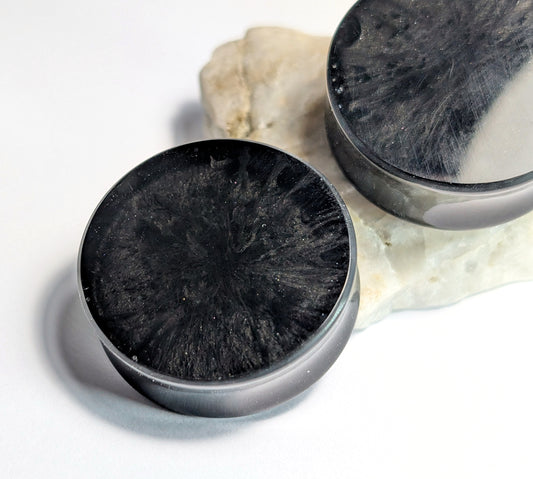 The Cerulean Wolfs Black Depths Double Flared Plugs, crafted from polished black mineral and featuring striking radiating patterns, are displayed on white stone, with a second plug partially visible against a white background.