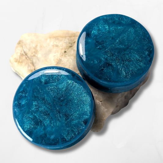 Two round, shiny Blue Ocean Double Flared Plugs by The Cerulean Wolf, featuring a marbled blue shimmer, are displayed on light textured stone with a plain white background.