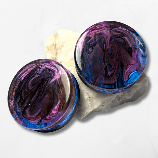 The Cerulean Wolfs Midnight Mist Double Flared Plugs, featuring glossy swirls of purple, blue, pink, and black, rest on a light-colored stone against a white background.