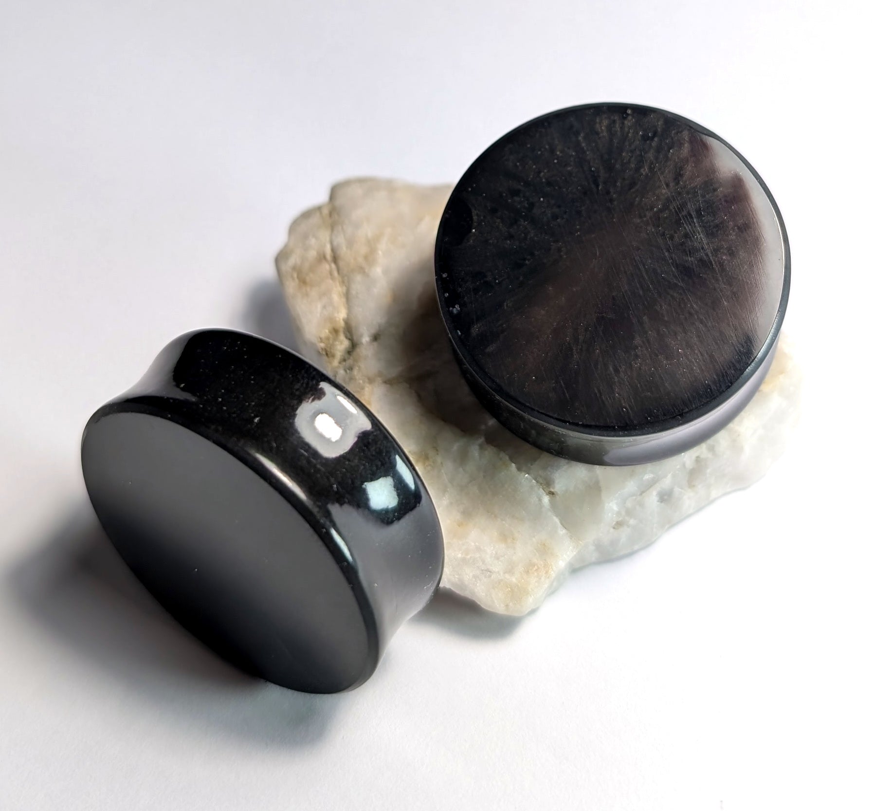 Two round Black Depths Double Flared Plugs by The Cerulean Wolf, with a polished, glossy finish and reflective surfaces, rest on a rough white stone surface against a clean white background.