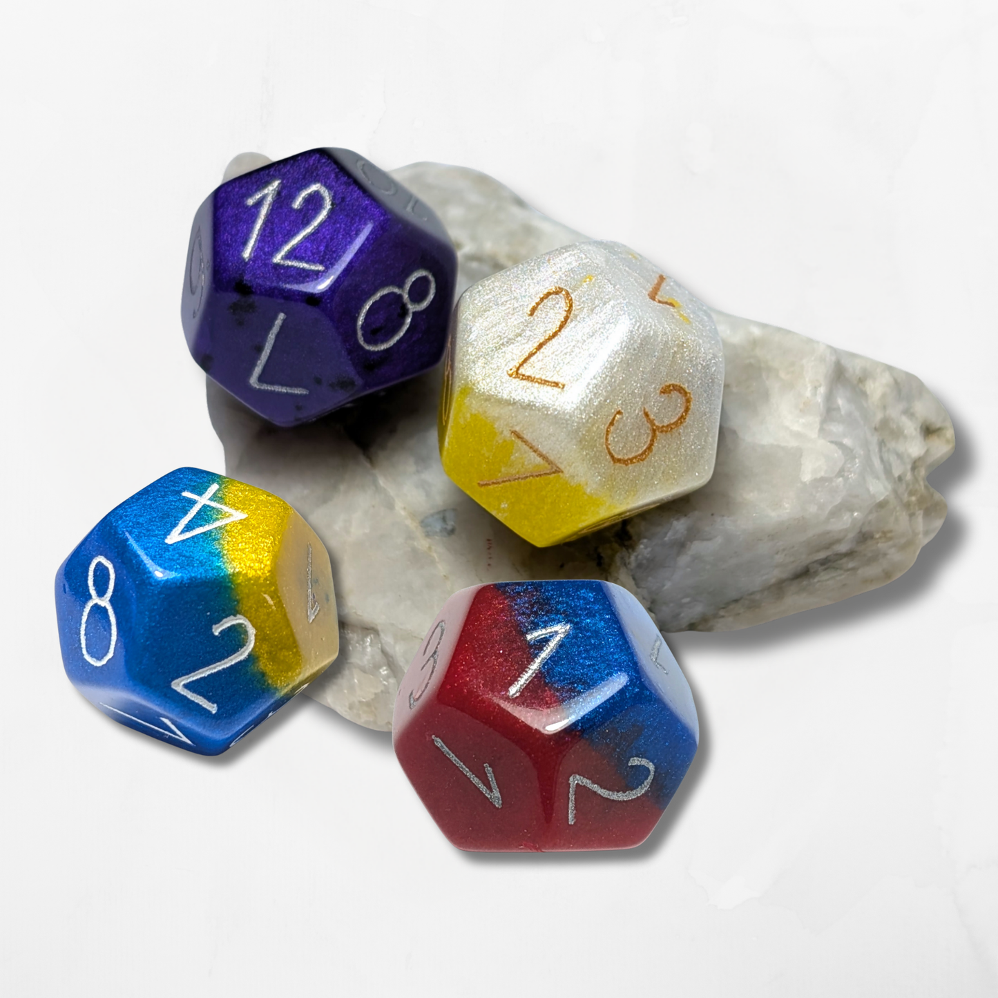 Four vibrant Custom Numbers & Colours D12 Dice by The Cerulean Wolf rest on white stone. Hand engraved in blue-yellow, red-blue, gold-white, and purple, these premium epoxy resin dice feature contrasting numbers for standout clarity.