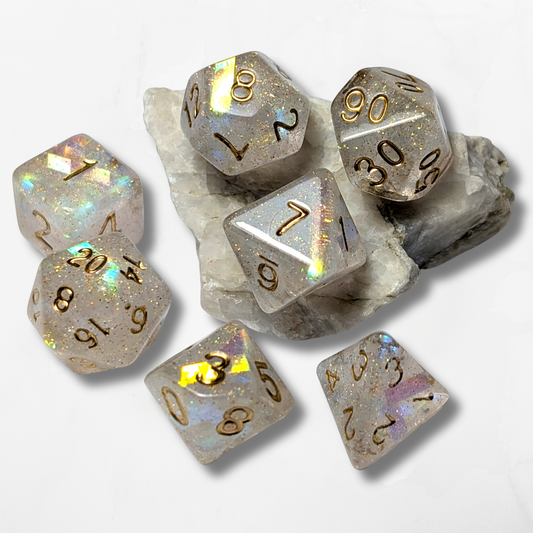The Cerulean Wolf’s 7 Holographic DND Dice, featuring glittery translucent resin and gold numbers, are displayed on and around a white and gray crystal stone against a white background.